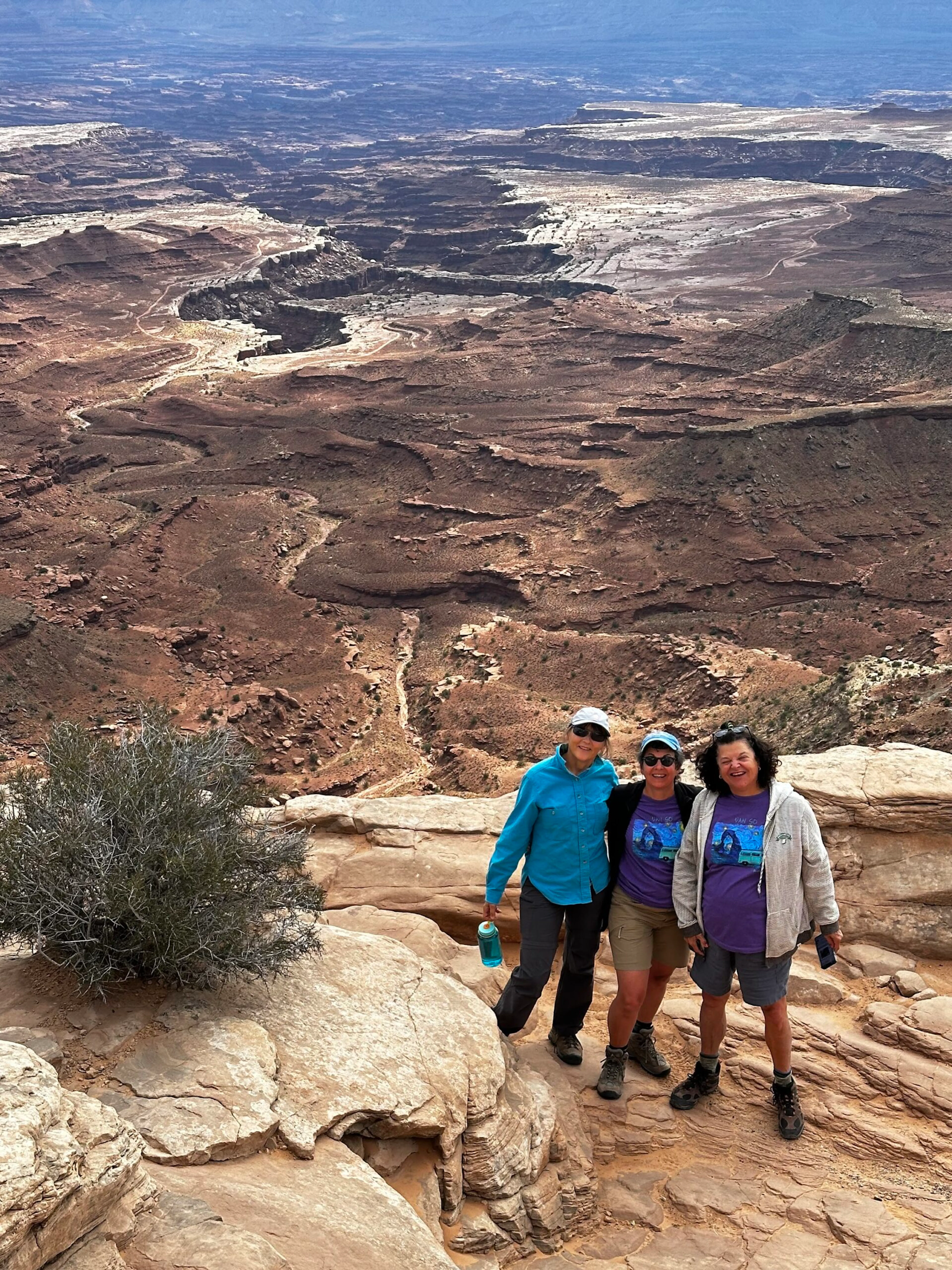 Three people pose on a rocky ledge overlooking a vast, rugged desert landscape with winding canyons—perfect for hiking and exploring the stunning scenery of Utah national parks.