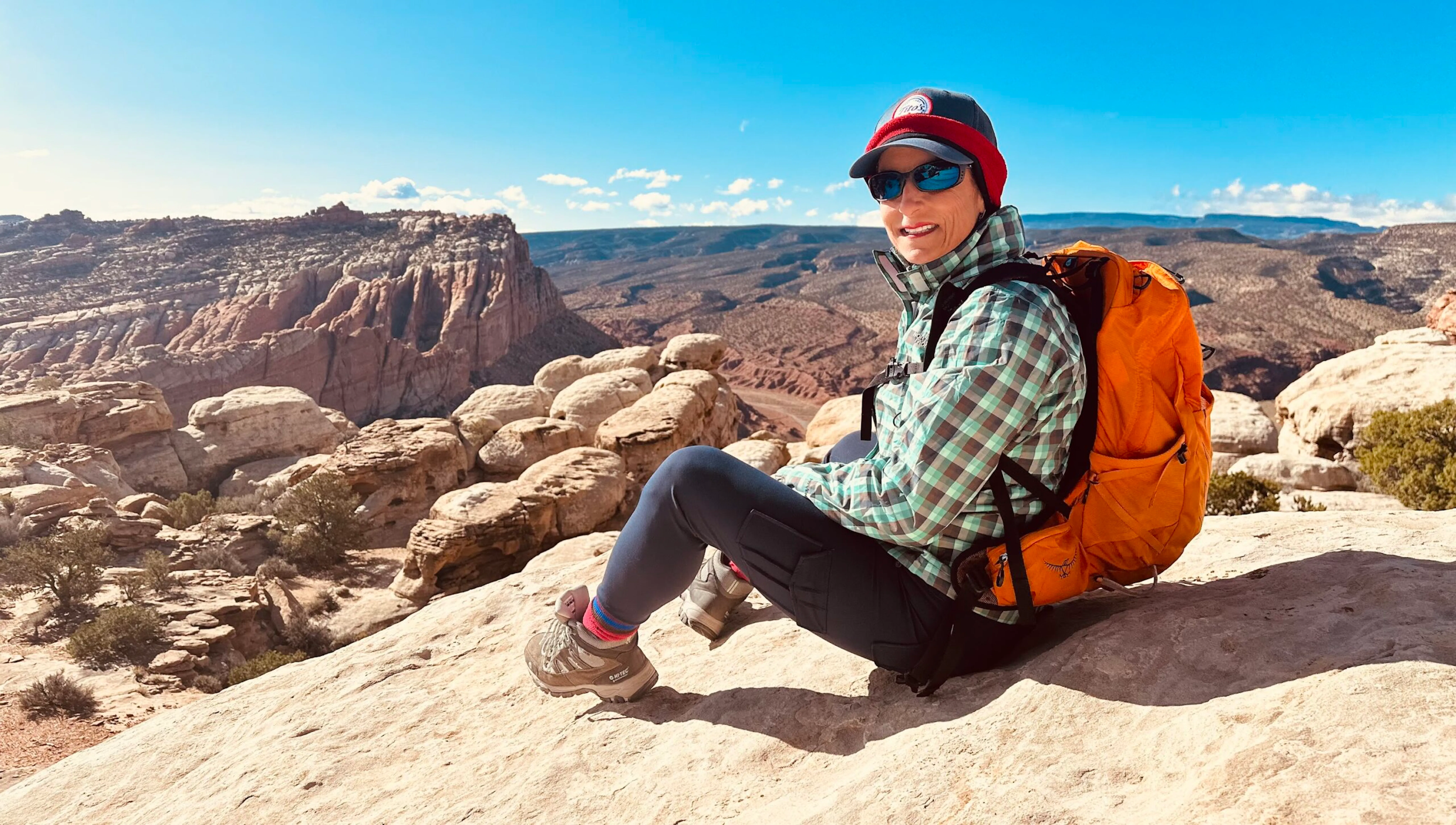 Person wearing outdoor gear and an orange backpack sits on a rocky ledge overlooking a desert canyon landscape in Utah national parks under a clear blue sky, capturing the spirit of adventure and hiking.
