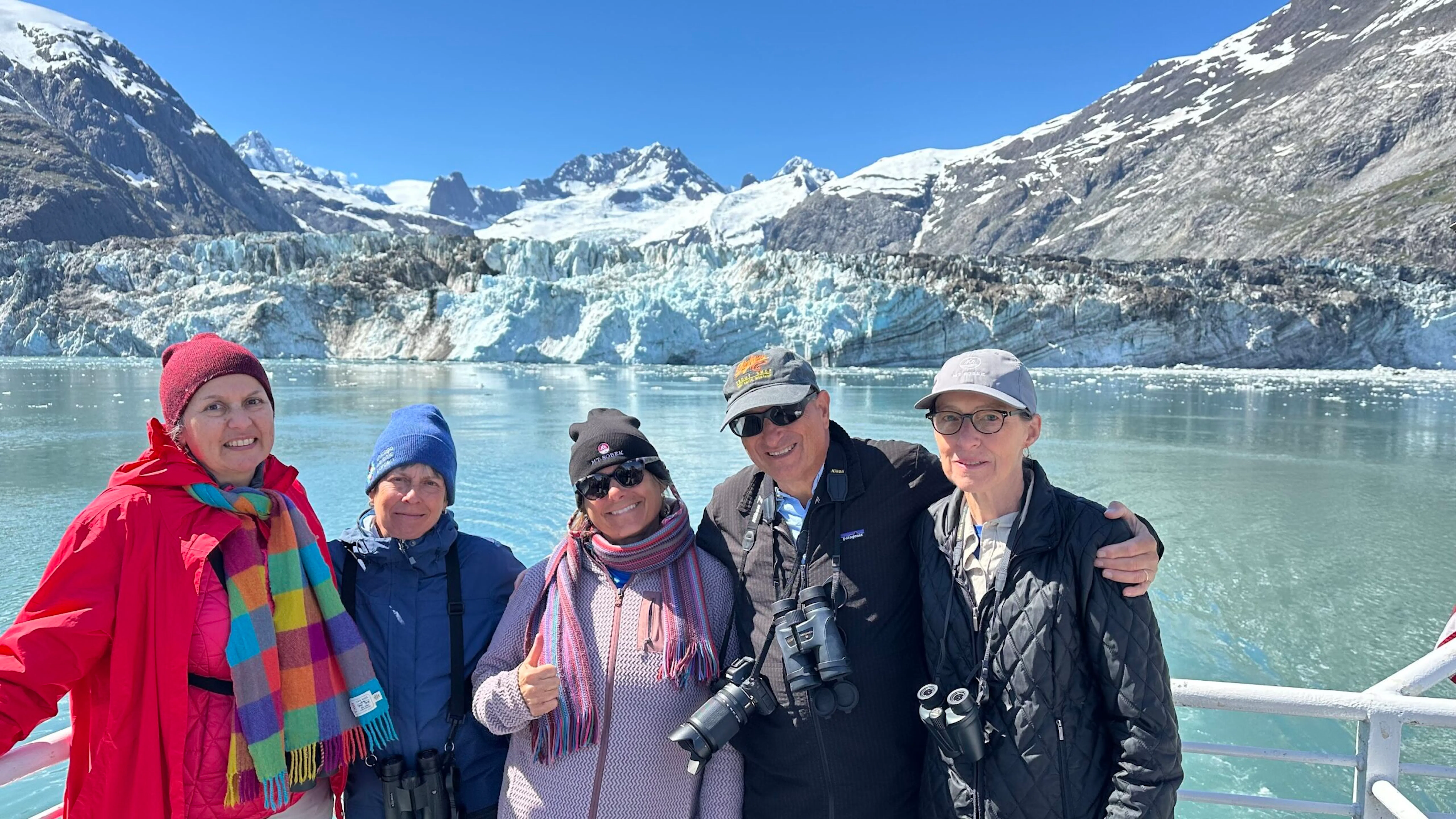Five people in outdoor clothing stand on a boat during one of their Alaska tours, with snowy mountains and a glacier in the background, smiling at the camera.