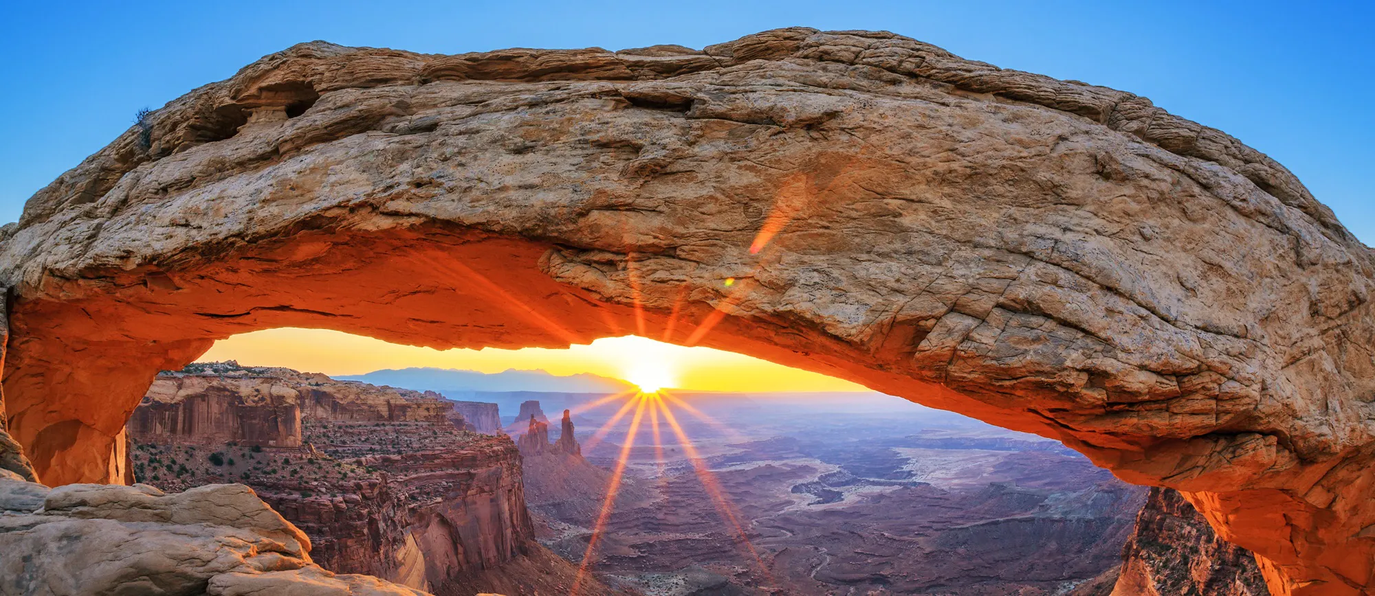 Sunrise shines through Mesa Arch, illuminating the rocky canyon landscape below—an unforgettable moment for hiking enthusiasts in Utah national parks.