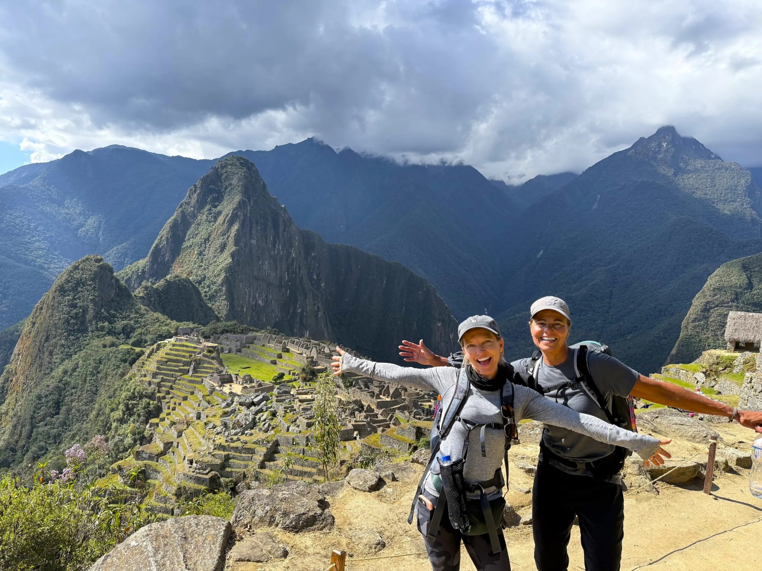 Two hikers posing and smiling with arms outstretched in front of the ancient Inca ruins of Machu Picchu, surrounded by mountains and cloudy sky.
