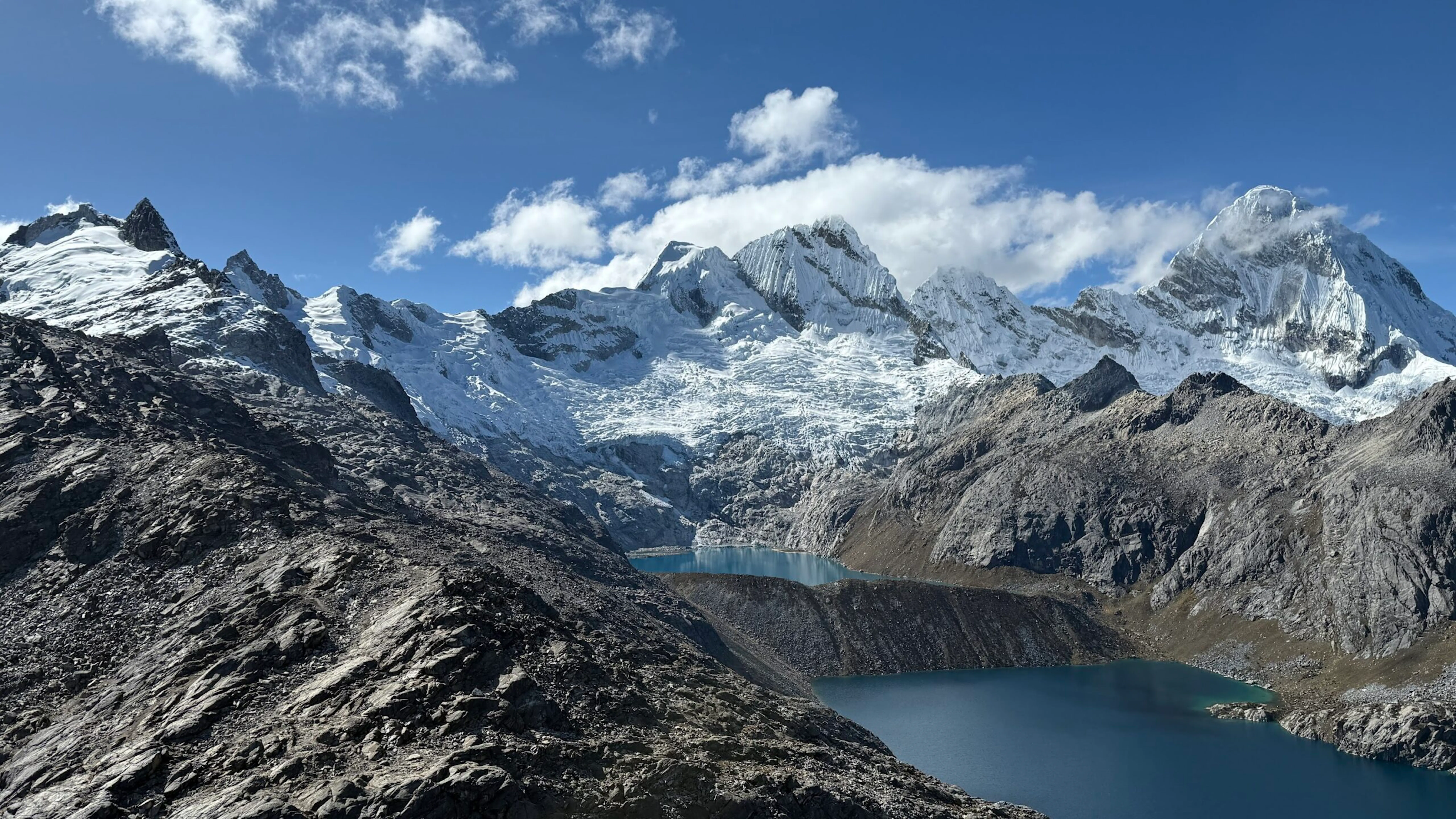 Snow-capped mountains rise above two blue lakes surrounded by rocky terrain under a mostly clear sky with scattered clouds.