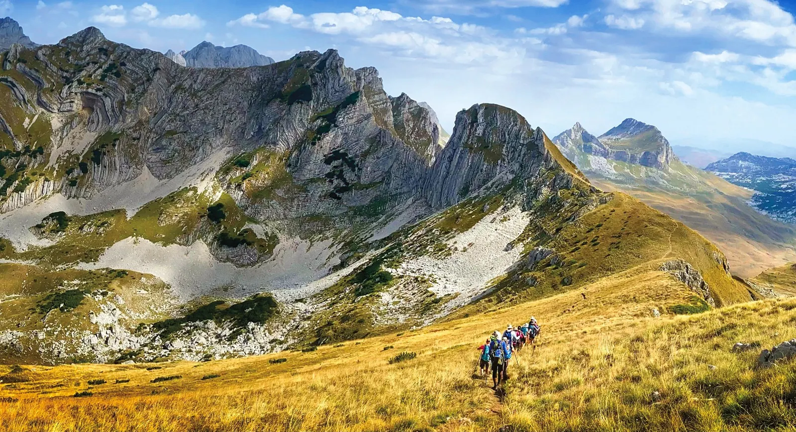 A group of hikers walks along a grassy mountain slope with rocky peaks and a partly cloudy sky in the background, embodying the spirit of adventure travel that draws explorers to breathtaking landscapes.