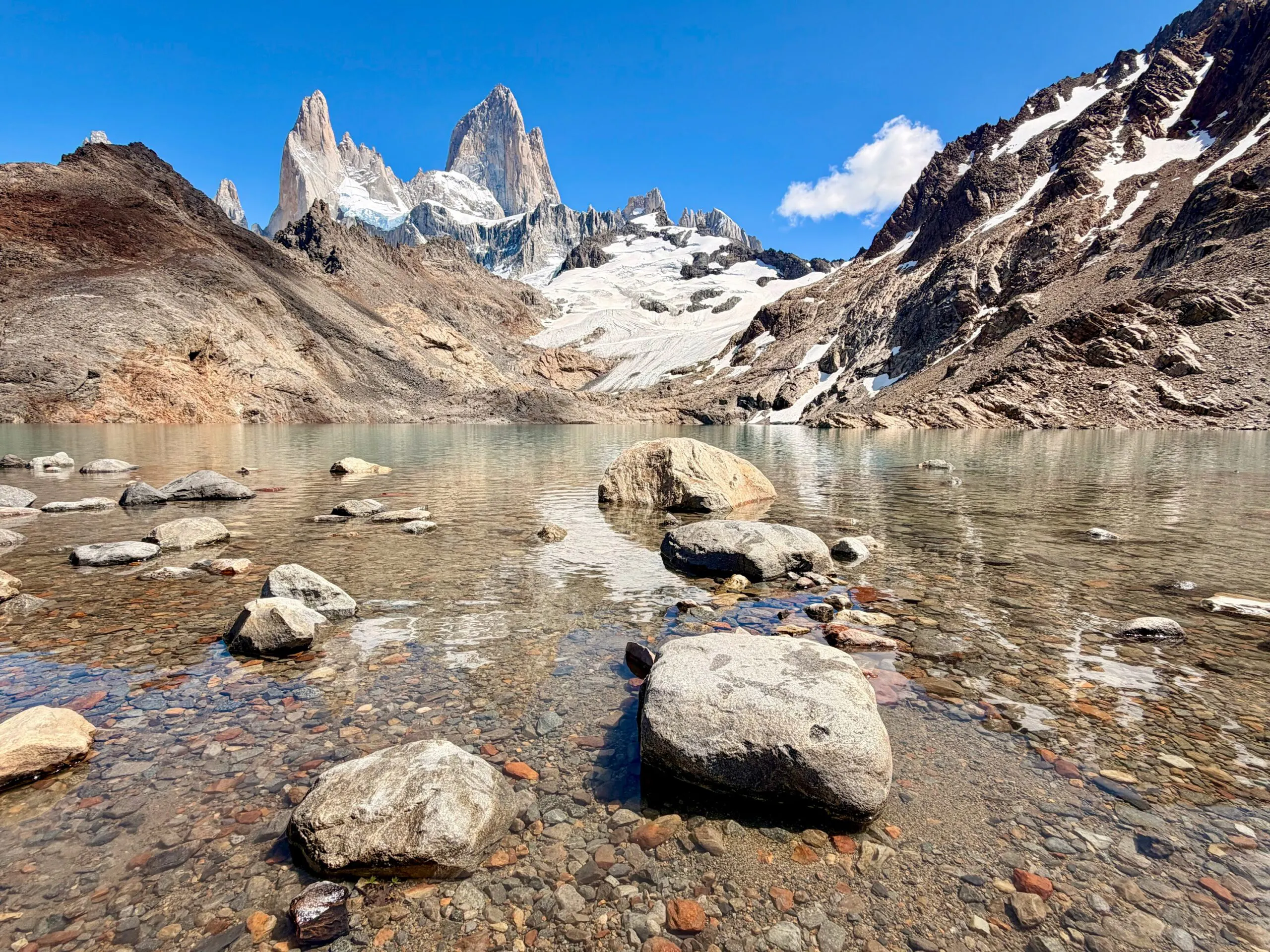 Patagonia clear glacial lake with rocks in the foreground, surrounded by rugged mountains and snow-capped peaks under a blue sky—a breathtaking spot for popular adventures.
