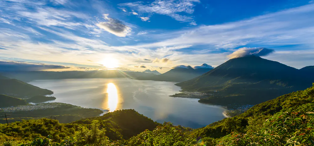 View of a large lake surrounded by green hills and mountains at sunrise, with sunlight reflecting on the water under a blue sky with scattered clouds.