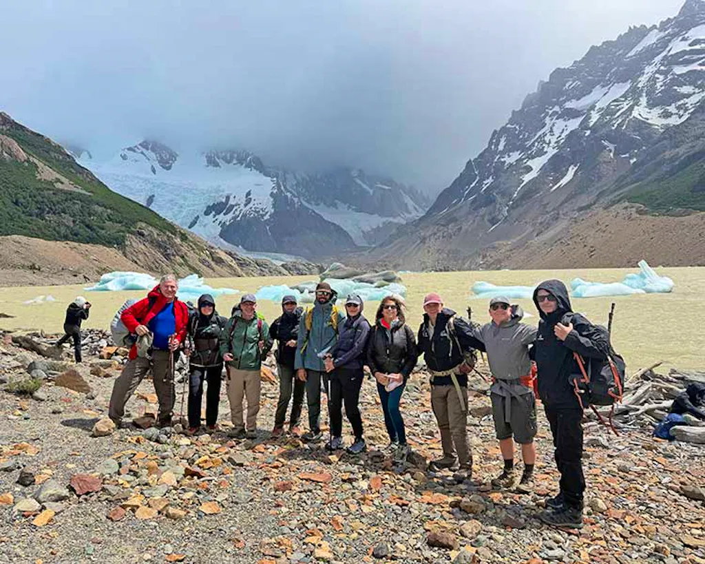 A group of hikers poses together on rocky terrain by a glacial lake in Argentina, with mountains and icebergs visible in the background under cloudy skies.