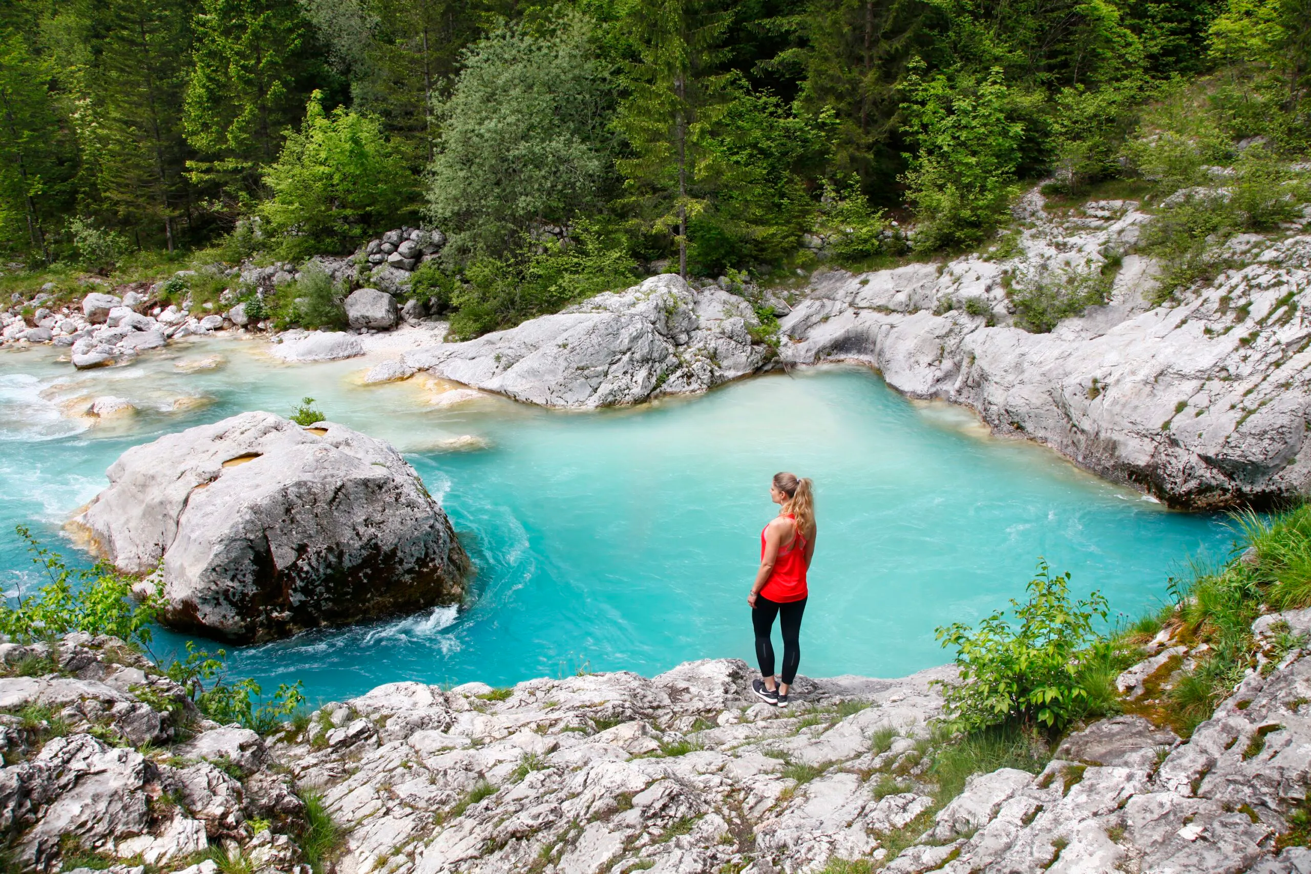 A person in a red top stands on a rocky bank, looking at a turquoise river surrounded by trees and rocks in a lush forest, as if patiently waiting their turn—like being on a waitlist for nature’s serene beauty.