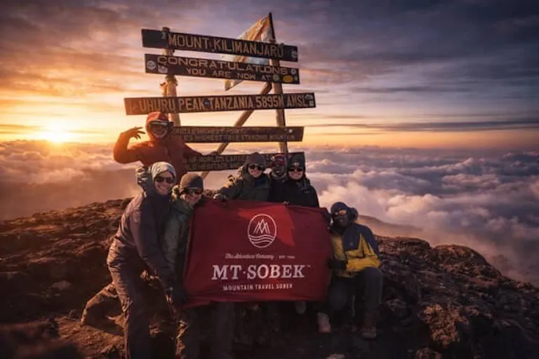 Six people pose at Mount Kilimanjaro’s summit with a Mt. Sobek banner in front of summit signs at sunrise, above the clouds.