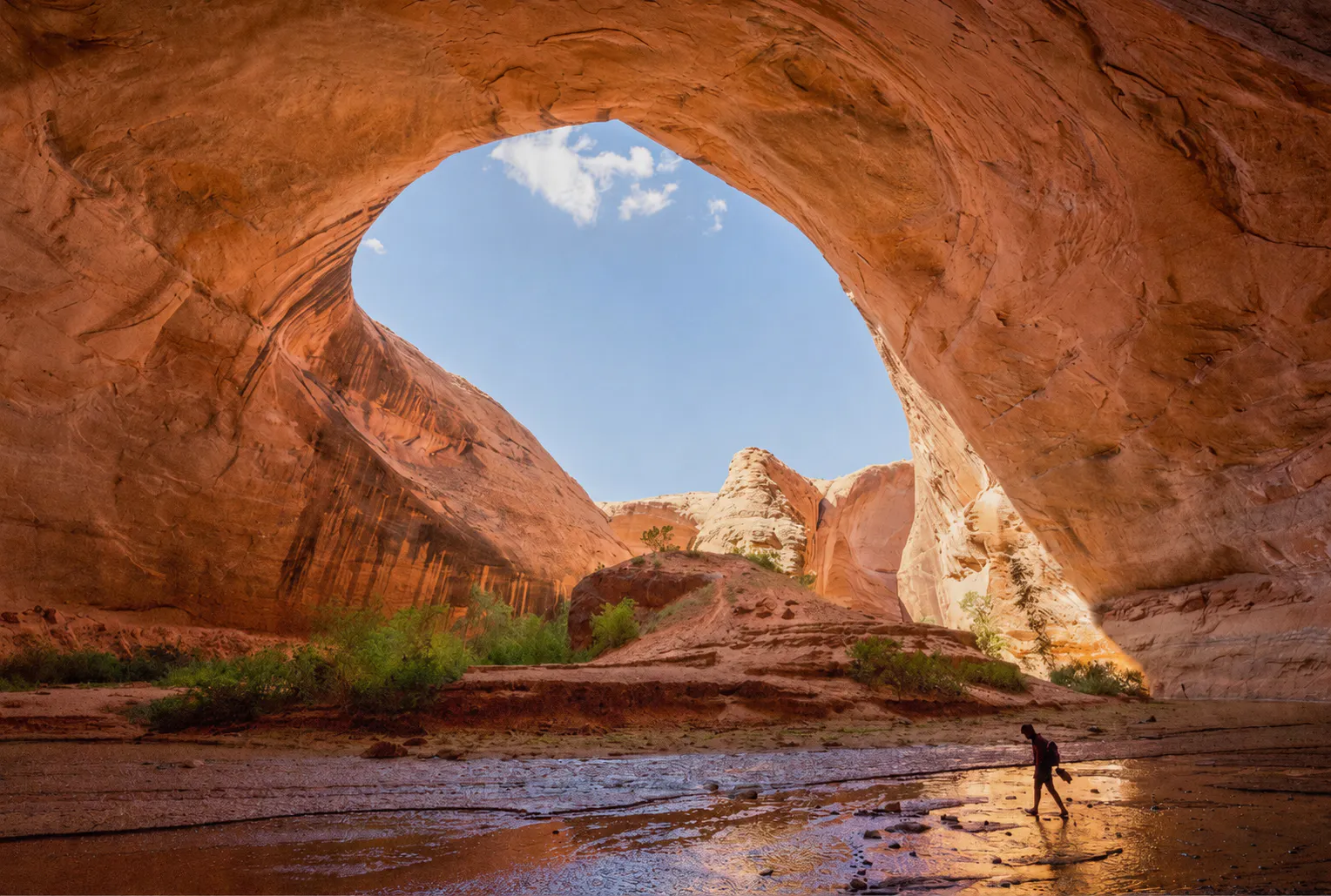 A person walks through shallow water inside a large red rock arch with sunlight illuminating the cave and blue sky visible above.
