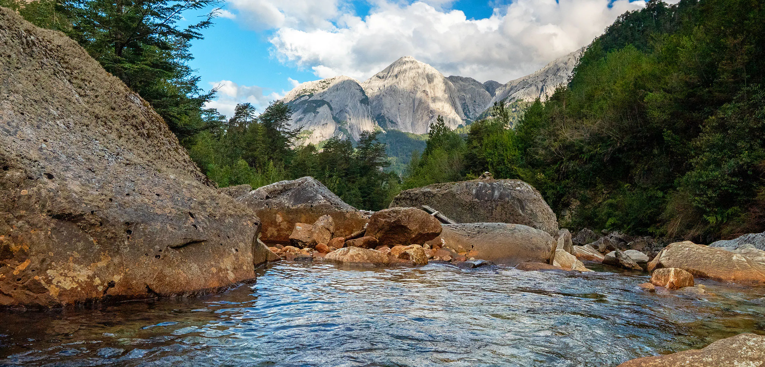 Clear stream flows over rocks, surrounded by green trees, with tall, snow-capped mountains and a partly cloudy blue sky in the background.
