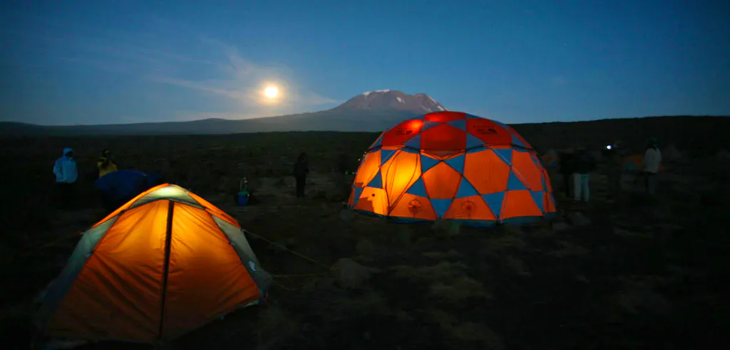 tents glowing at night with hikers starting around, and the full moon and Kilimanjaro in the background