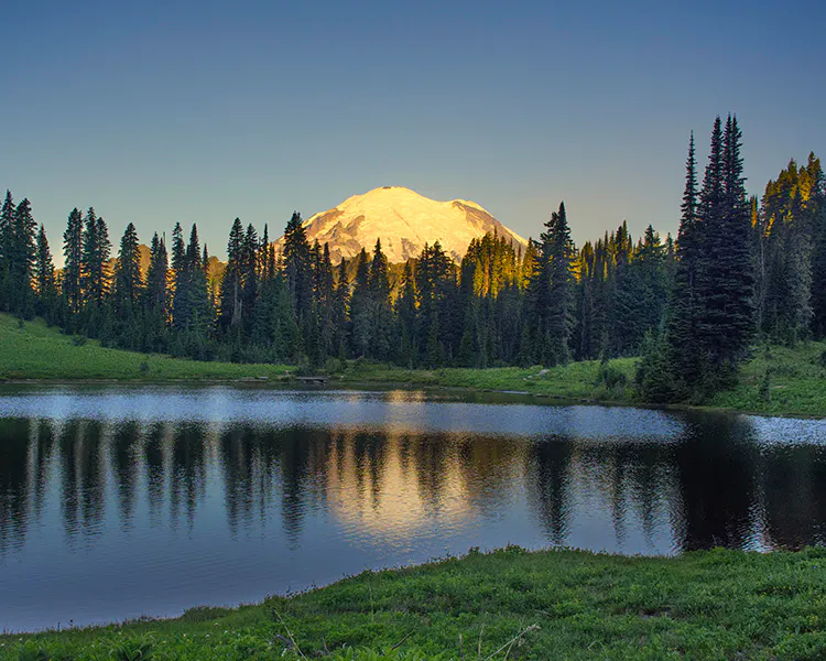 Mountain lake ringed by conifer forests with Mount Rianier in the background at sunset