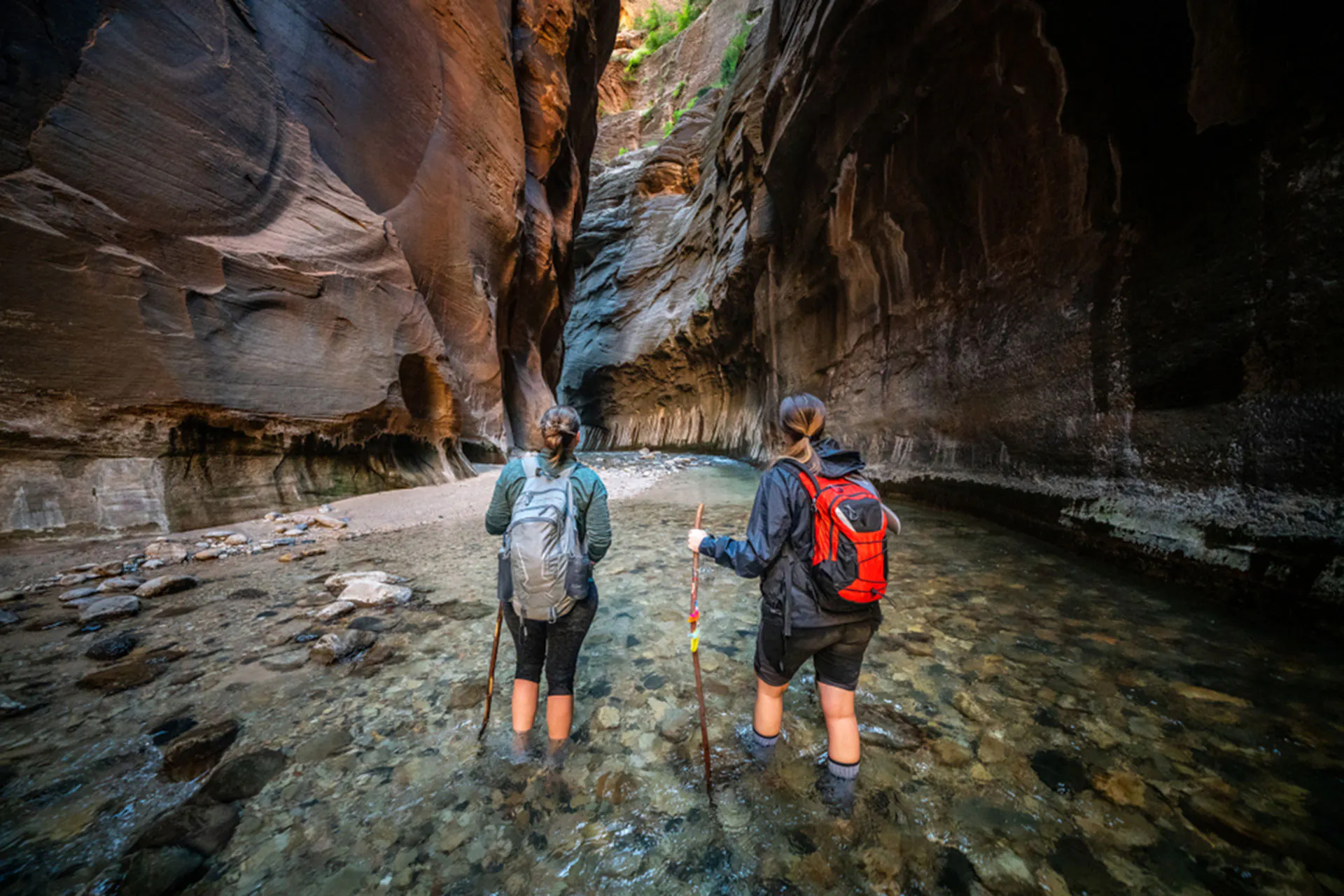 Two female hikers in the Zion Virgin River Narrows, standing on river rocks in the water looking up at sun reflecting off sandstone walls