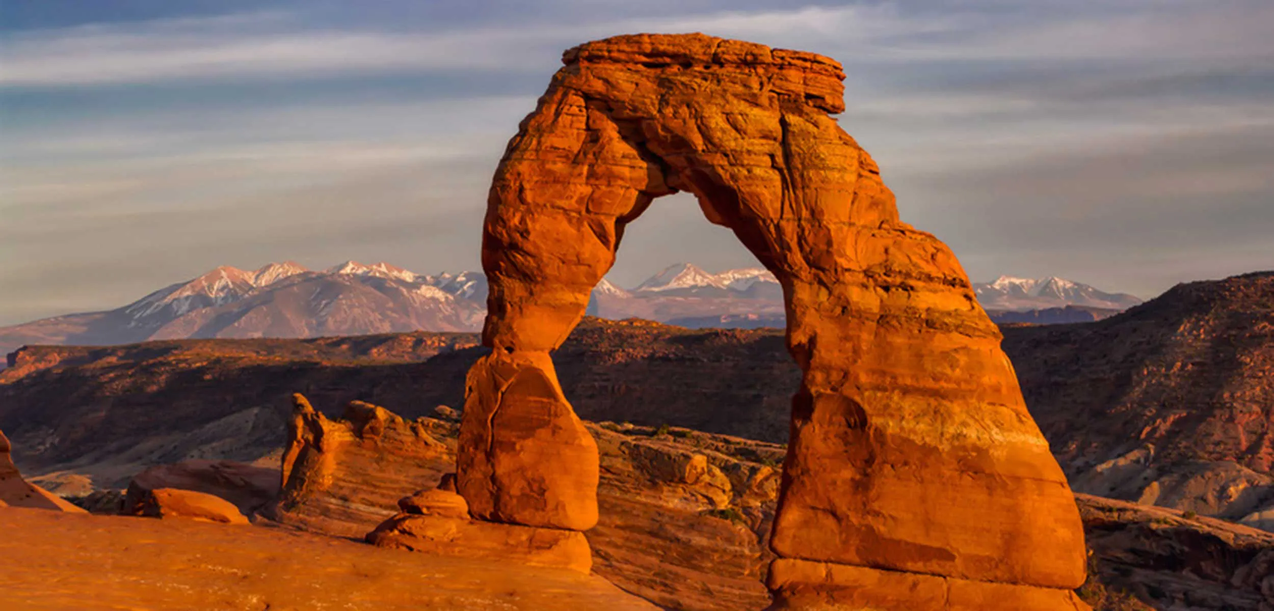 View of Delicate Arch in Arches National Park at sunset with the snow-capped La Sal Mountains in the background