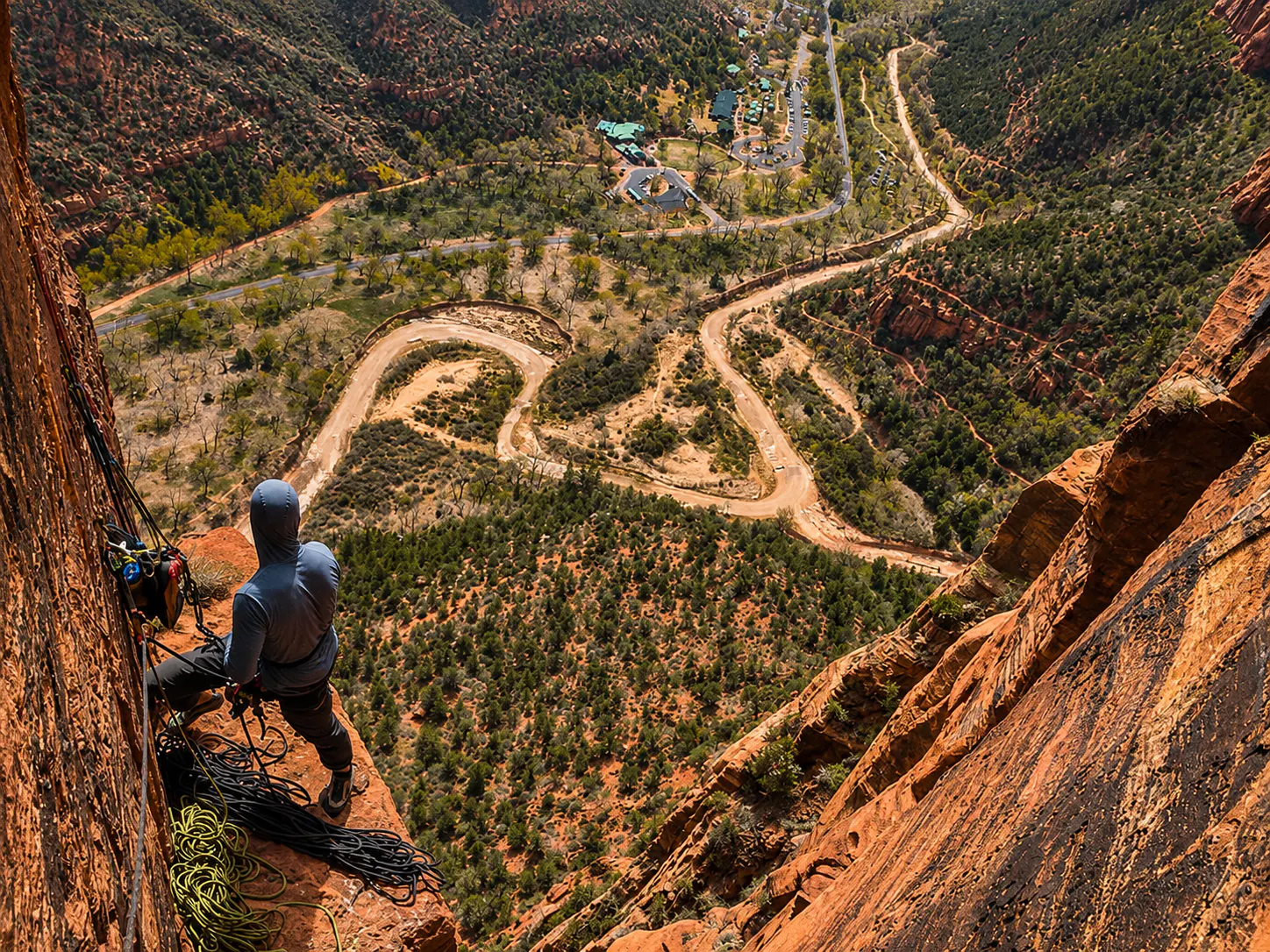 A climber in Zion National Park in a blue sun hoody looking down from a belay ledge at the Virgin River and Zion Canyon