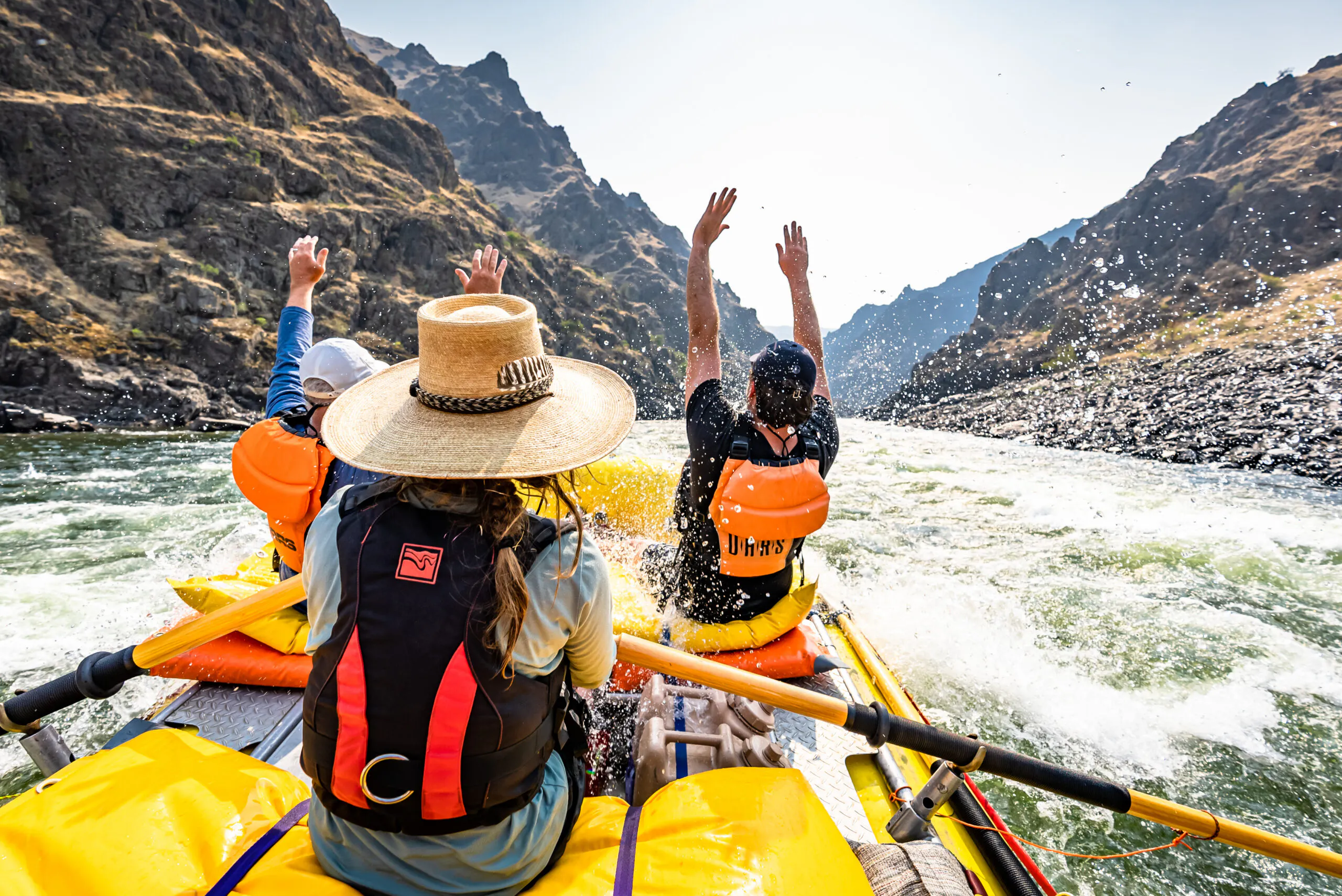 Three people in life jackets ride a raft down a river between rocky cliffs, with water splashing and two people raising their arms in excitement.