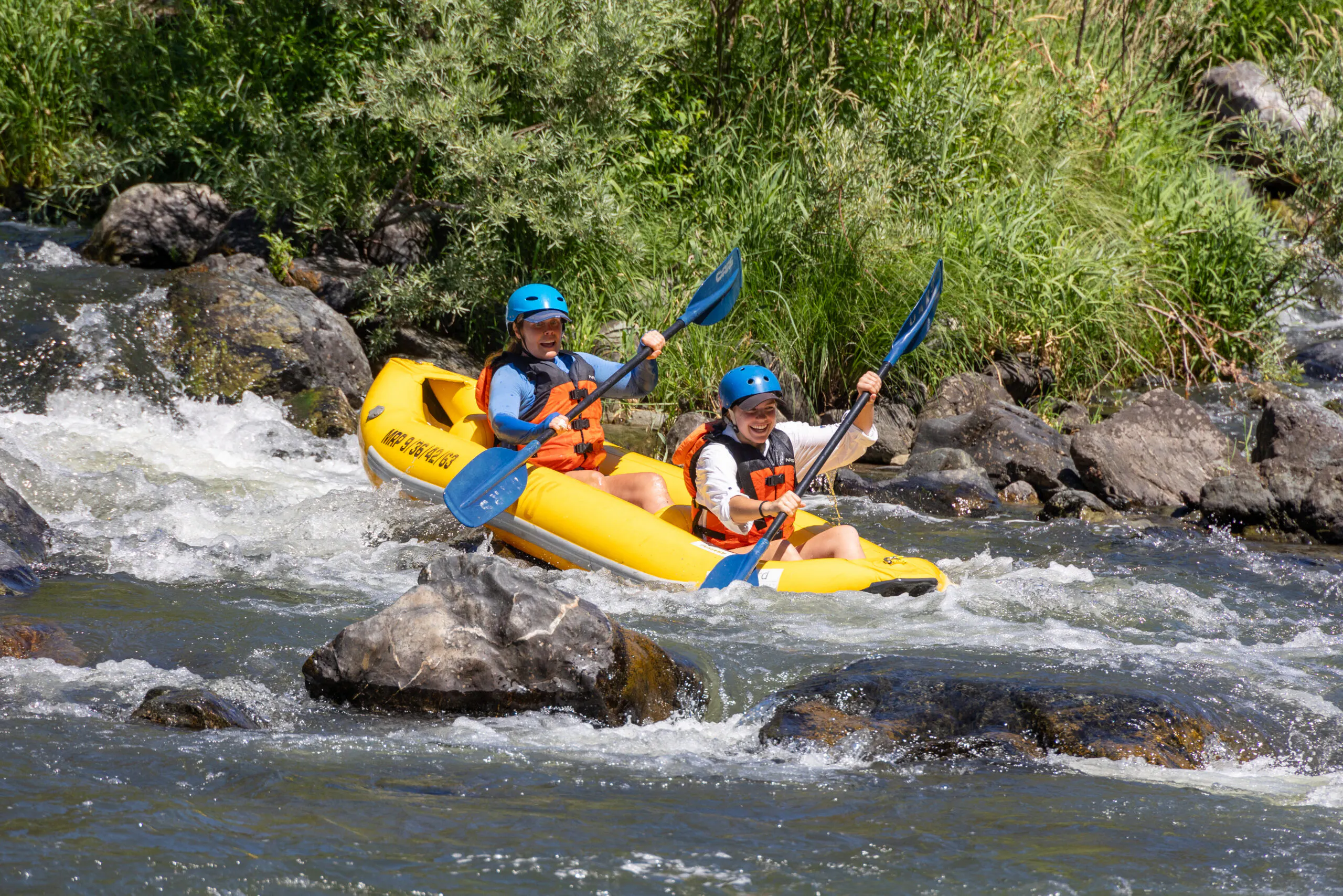 Two people wearing helmets and life jackets paddle a yellow inflatable kayak through whitewater rapids surrounded by rocks and greenery.