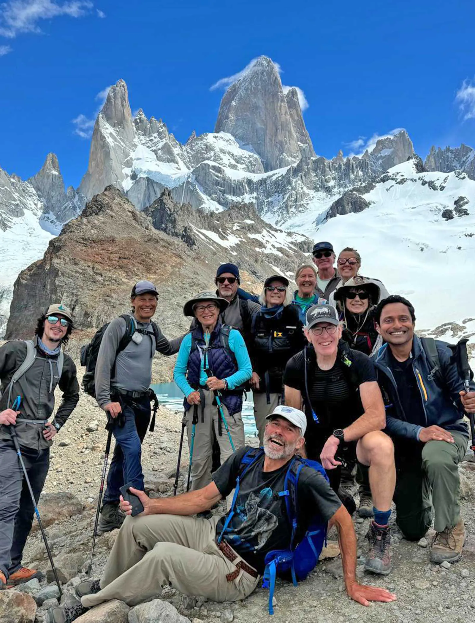 A group of hikers pose on rocky terrain with trekking poles, smiling in front of snowy mountains under a clear blue sky—a perfect snapshot from one of our unforgettable South America adventure tours.