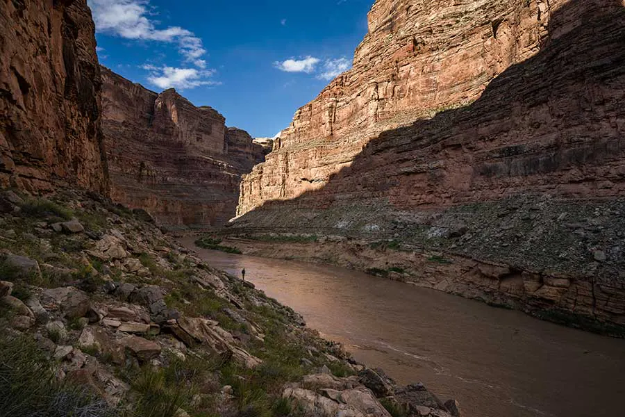 The Colorado River flowing through Cataract Canyon, with narrow, towering walls and a man walking along shore in the distance—OARS trip in Utah