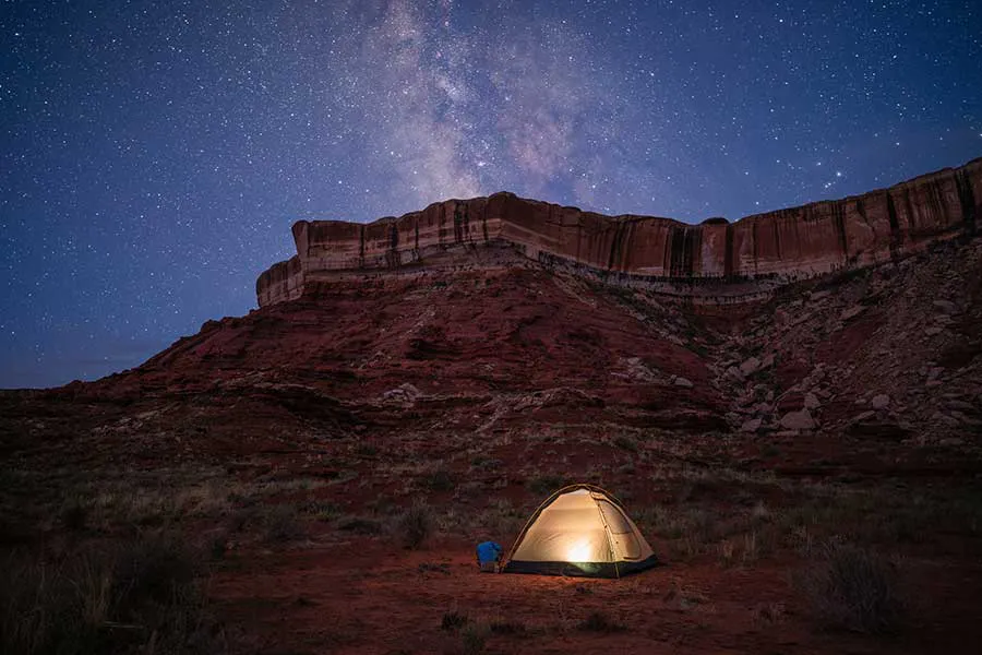A guest's tent on an OARS trip in Cataract Canyon; tent lit up by a light and a man sitting outside looking up at sandstone cliffs and the Milky Way