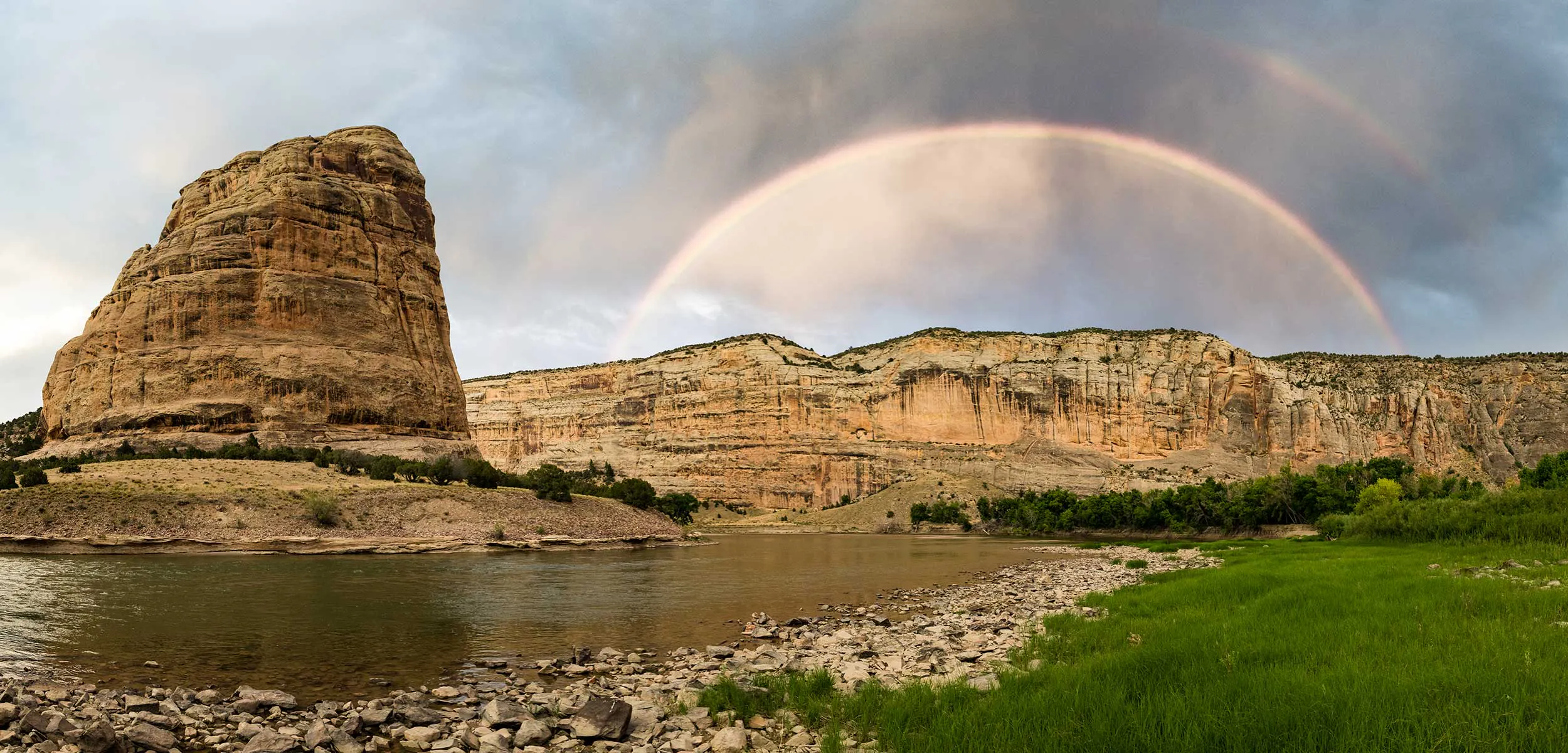 A rocky cliff and riverbank under a cloudy sky with a double rainbow arching across the background.