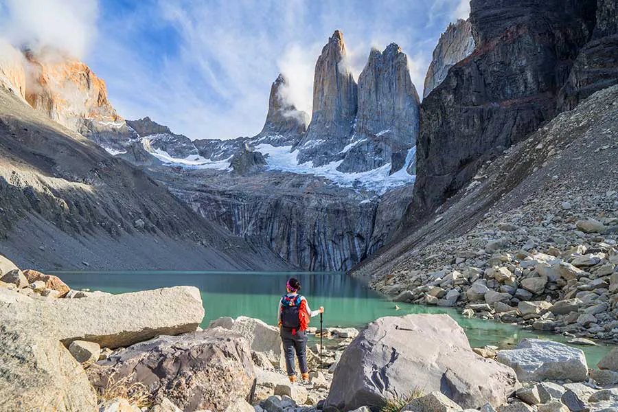 A person with a red backpack stands on rocky terrain, immersed in Patagonia hiking, facing a turquoise lake and dramatic mountain peaks under a partly cloudy sky.