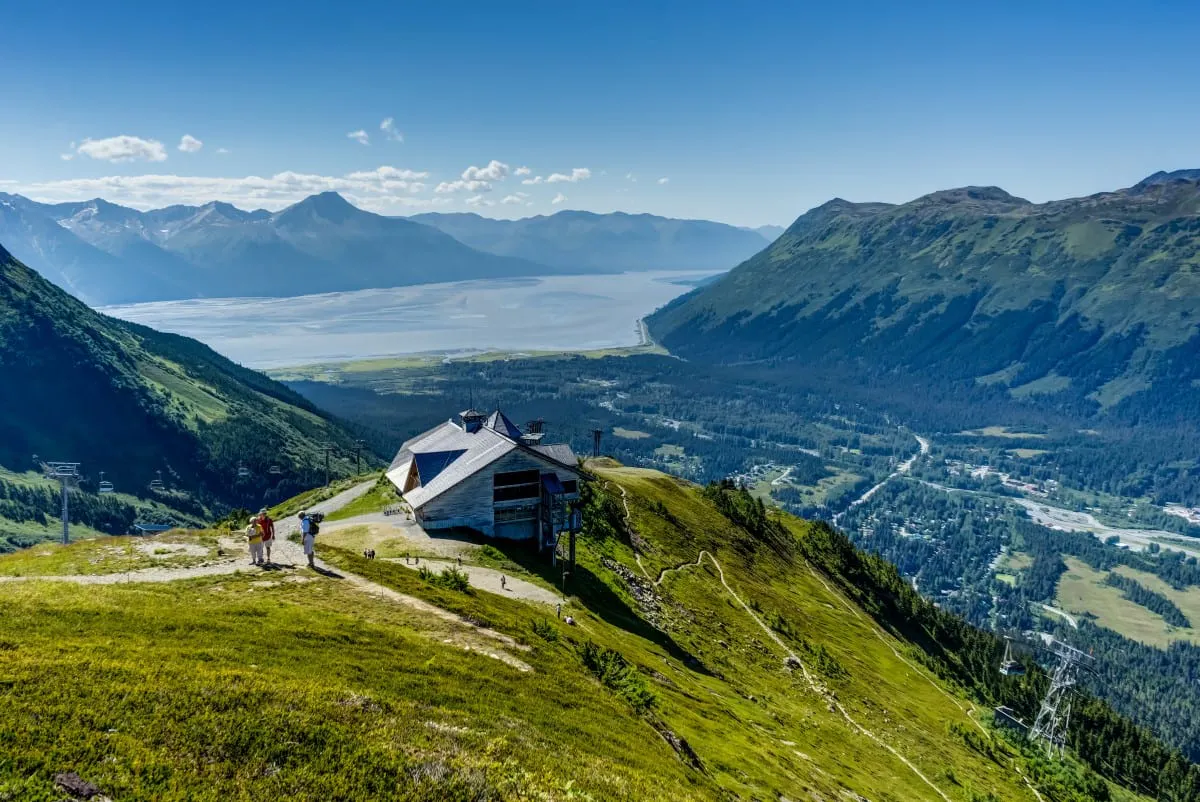 Alyeska Ski Resort in Alaska sits on a green mountainside overlooking a valley, river, and distant mountains under a clear blue sky—the perfect spot to pause and take in the breathtaking views after a day of hiking.