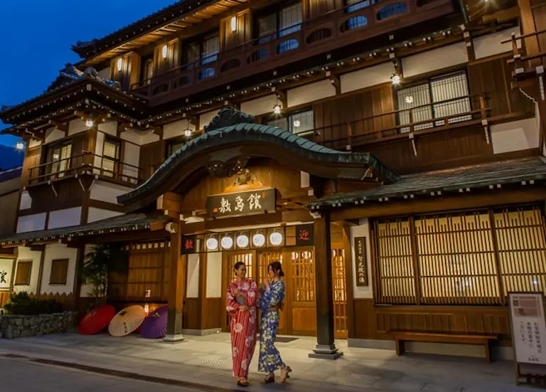 Two people in traditional kimonos stand in front of a lit-up wooden Japanese building at dusk, their colorful umbrellas placed beside the entrance of Onyado Shikishimakan in Japan.