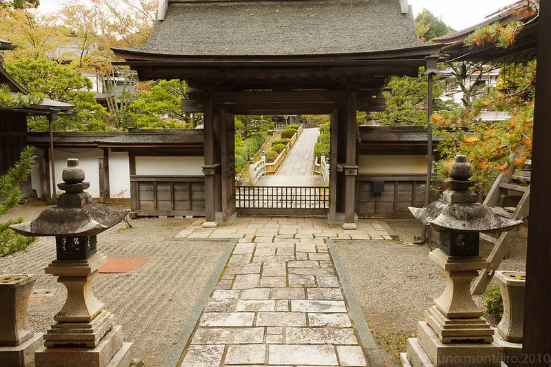 Stone pathway leading through a traditional Japanese gate at the Shojoshin-in, with stone lanterns on each side, surrounded by greenery and wooden buildings—perfect for a peaceful hiking experience amidst timeless scenery.