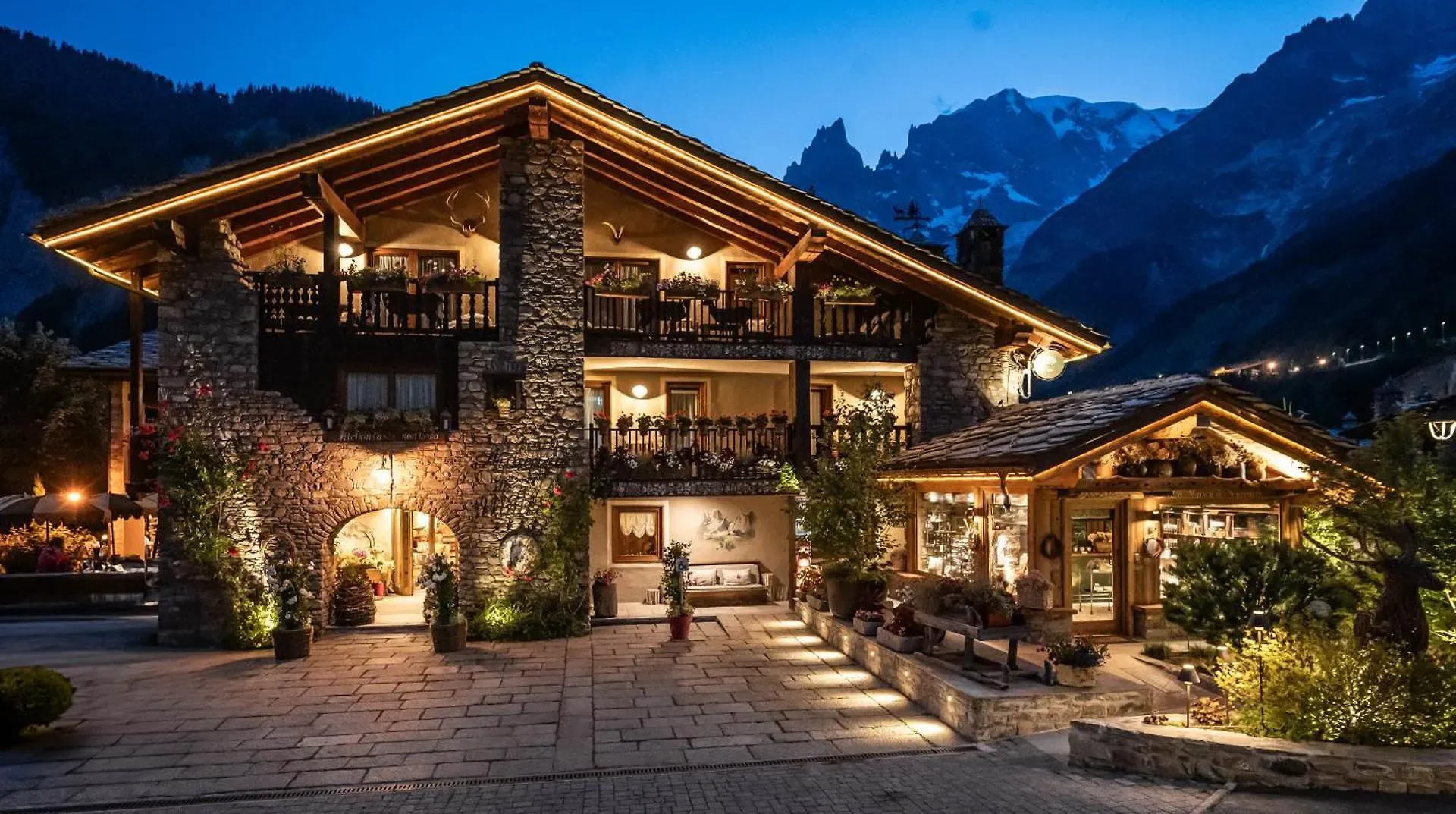 Auberge de la Maison with warm lights and wooden balconies stands in a mountain village at dusk, inviting hikers to rest after a day of hiking among the tall peaks visible in the background.