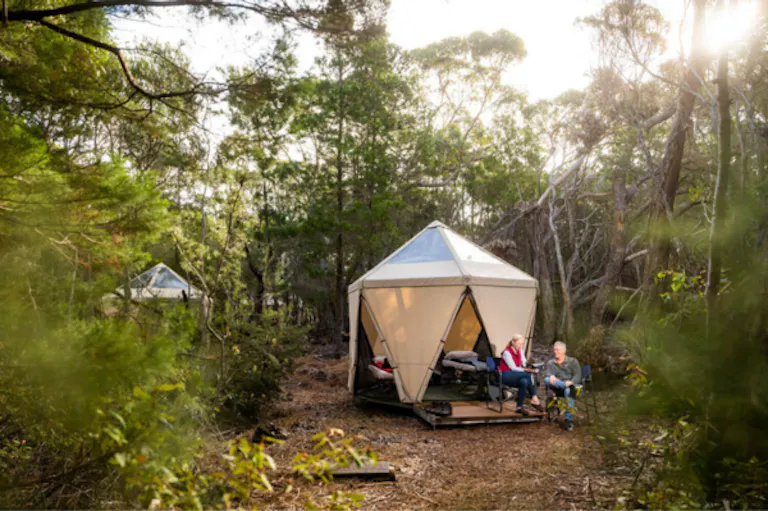 Two people sit outside a modern tent that belongs to Flinders Island Eco Camp, in a forested area, relaxing after a day of hiking, with another similar tent visible in the background. Sunlight filters through the trees.