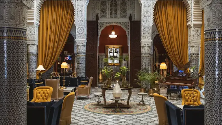 Ornate lounge of Riad Fes in Morocco with patterned columns, yellow drapes, mosaic floors, and plush seating, featuring elegant lamps and plants that evoke the serenity one might seek after a long day of hiking.