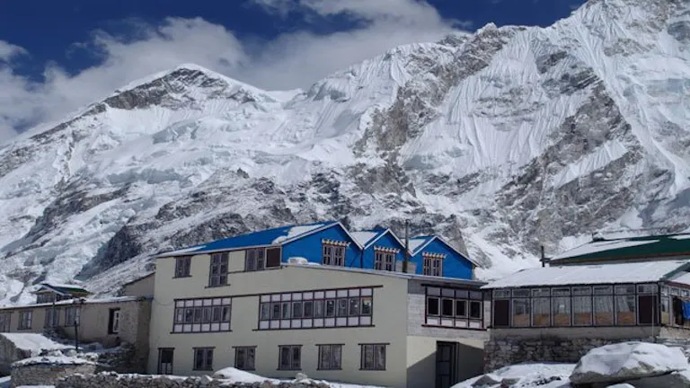 Snowland Highest Inn with blue roofs are set against a backdrop of rugged, icy mountains under a partly cloudy sky, creating an inviting scene for those passionate about hiking in pristine winter landscapes.