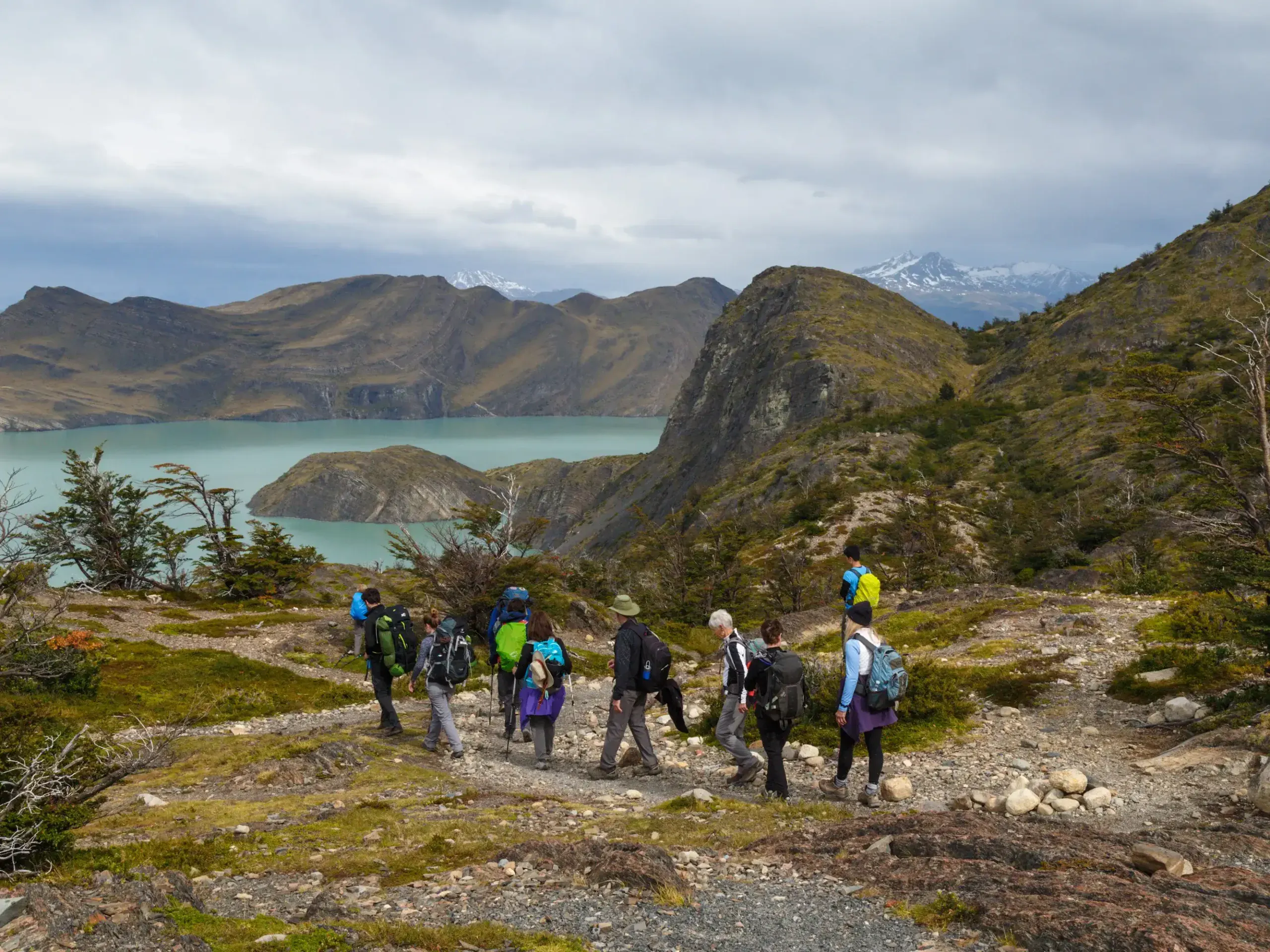 A group of hikers with backpacks walks along a rocky trail in Torres del Paine, overlooking a turquoise lake and mountains under a cloudy sky.