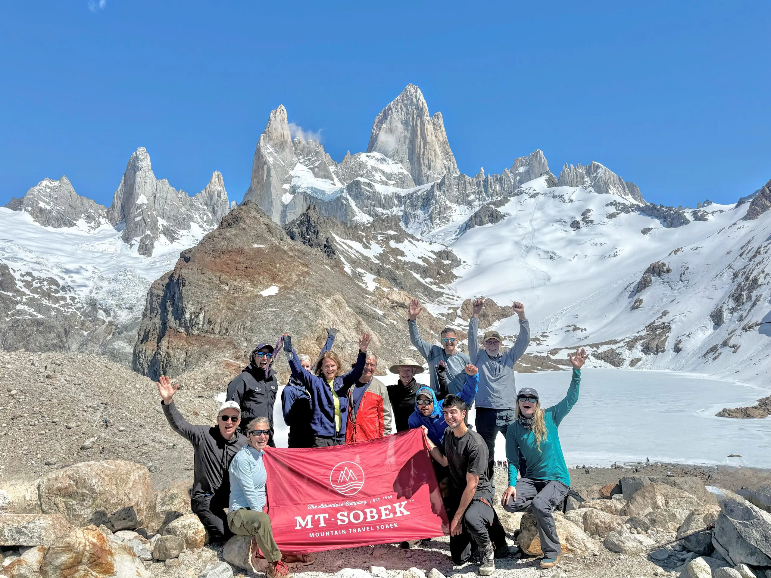 A group of people poses with a red “Mt. Sobek” banner in front of snow-covered mountains and rocky terrain under a clear blue sky, capturing the spirit of adventurous Patagonia hikes.