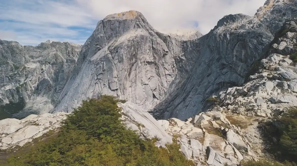 Steep, rugged granite cliffs rise behind green trees in Cochamo Valley, Patagonia under a partly cloudy sky in a mountainous landscape.