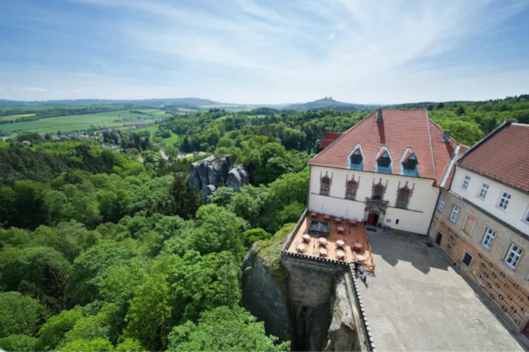 Historic building that belongs to Chateau Hruba Skala, with red roof overlooks a forested landscape and distant hills under a blue sky; terrace with tables visible in the foreground, making it an inviting spot to relax after a day of hiking nearby.