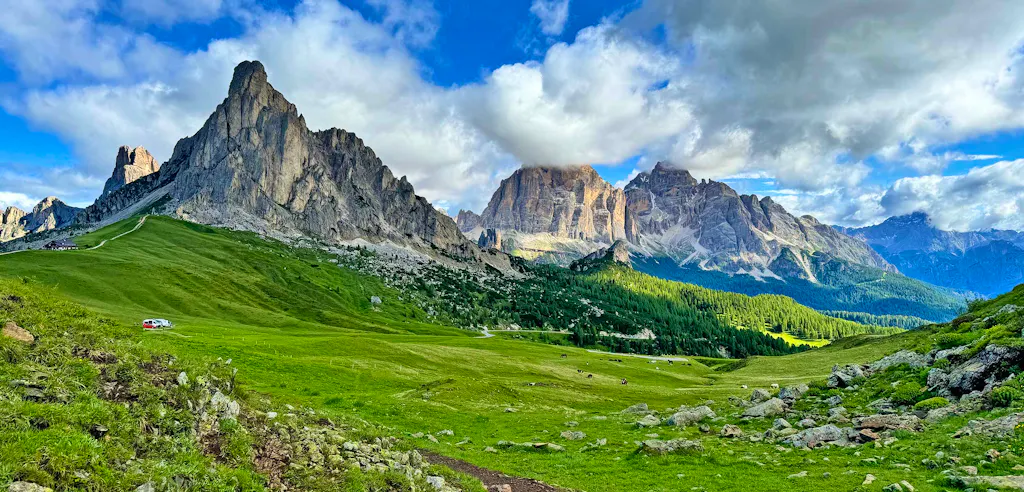 Jagged mountain peaks under a partly cloudy sky rise above a wide green meadow with scattered rocks and a single white vehicle visible on the left side, capturing the breathtaking scenery found along the best Dolomites hiking tours.