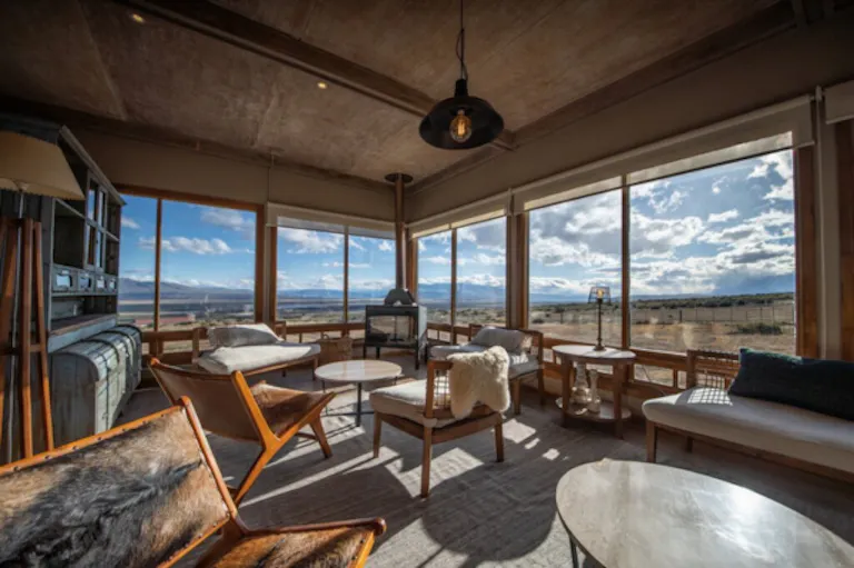 Estancia Cerro Guido's modern living room with large windows, wooden furniture, and views of open fields and mountains under a partly cloudy sky.