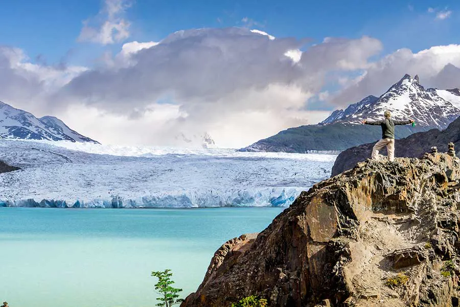 Person standing on a rocky outcrop with arms raised, overlooking a turquoise lake and a glacier surrounded by snow-capped mountains under a partly cloudy sky—a breathtaking scene typical of Patagonia hiking adventures.