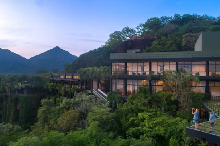 Heritance Kandalama Hotel in Sri Lanka, with large windows nestled in lush green hills, with mountains in the background and two people standing on a balcony