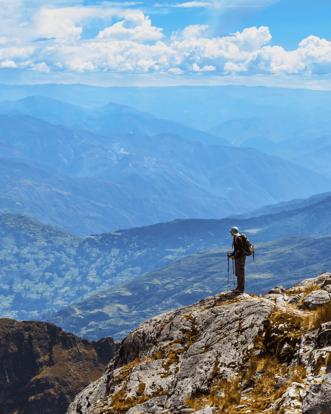 A person with hiking poles stands on a rocky cliff in Bolivia, overlooking layered mountain ranges under a partly cloudy sky, embodying the spirit of Bolivia hiking.