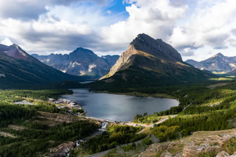 Glacier National Park landscape with a lake in the foreground, surrounded by dense green forest and Many Glacier Hotel near the water’s edge under a partly cloudy sky—an idyllic spot perfect for hiking enthusiasts to explore nature’s tranquility.