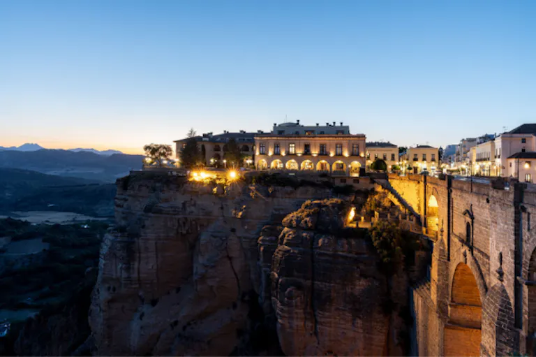 The Parador de Ronda in Spain with arched windows is illuminated at dusk atop a cliff, next to a historic stone bridge in a mountainous landscape, inviting hiking enthusiasts to explore the scenic trails nearby.