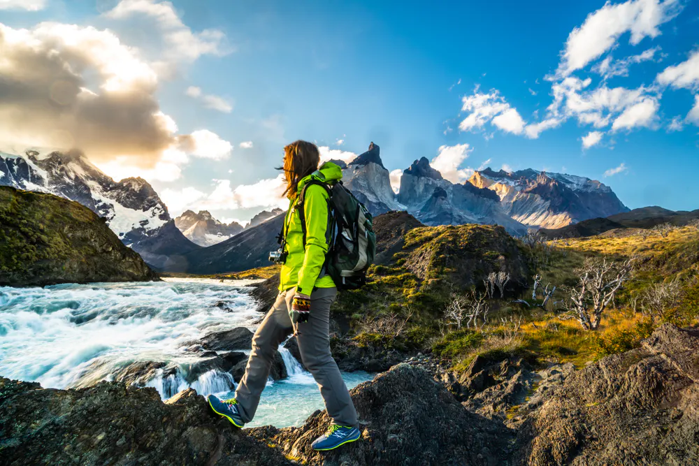 A person wearing hiking gear stands by a river with mountains and blue sky in the background on the W Trek in Torres del Paine National Park, Chilean Patagonia