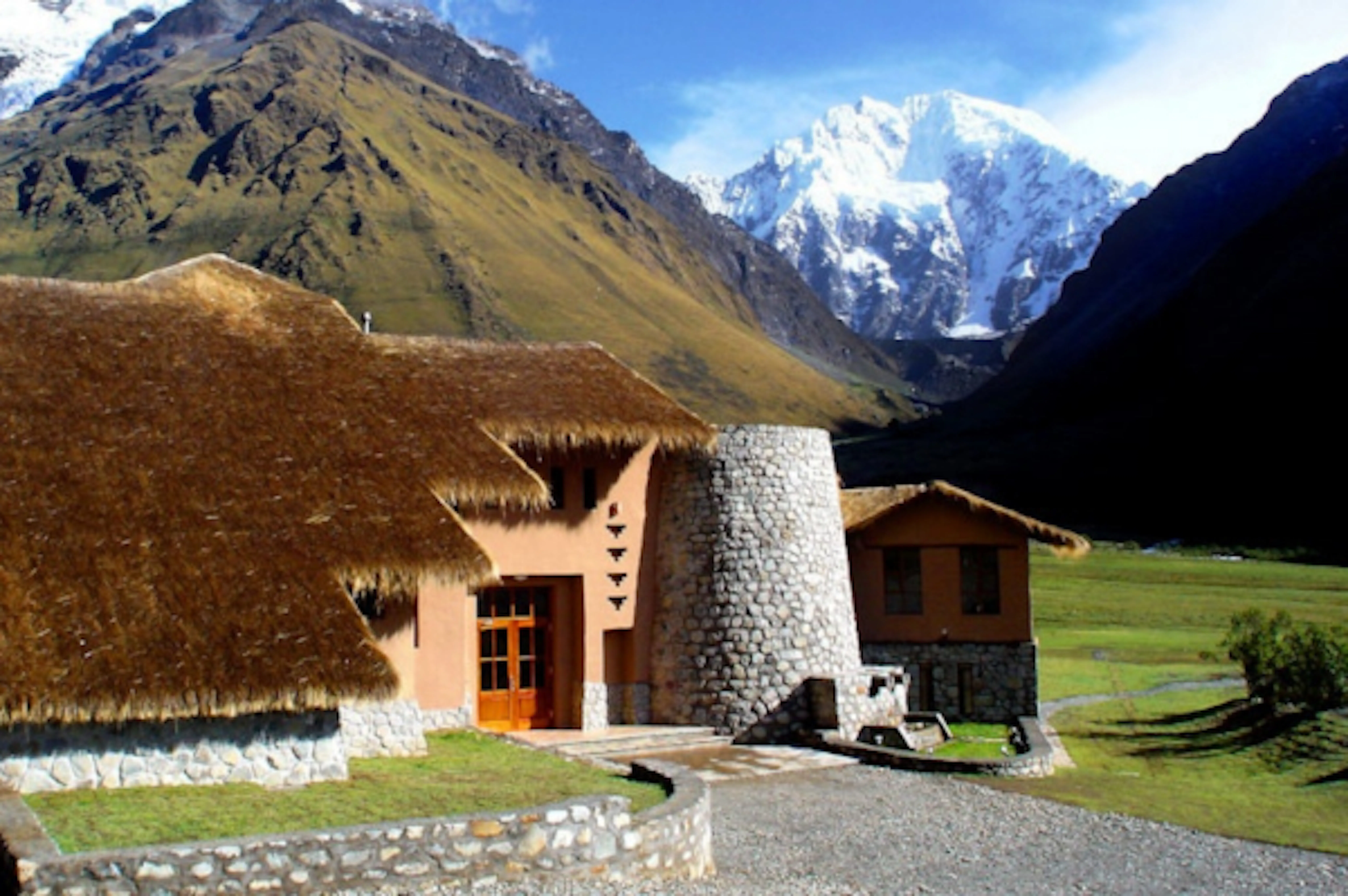 Salkantay Lodge with stone walls sit in the trail leading to Machu Picchu, surrounded by tall mountains with snow-covered peaks under a partly cloudy sky