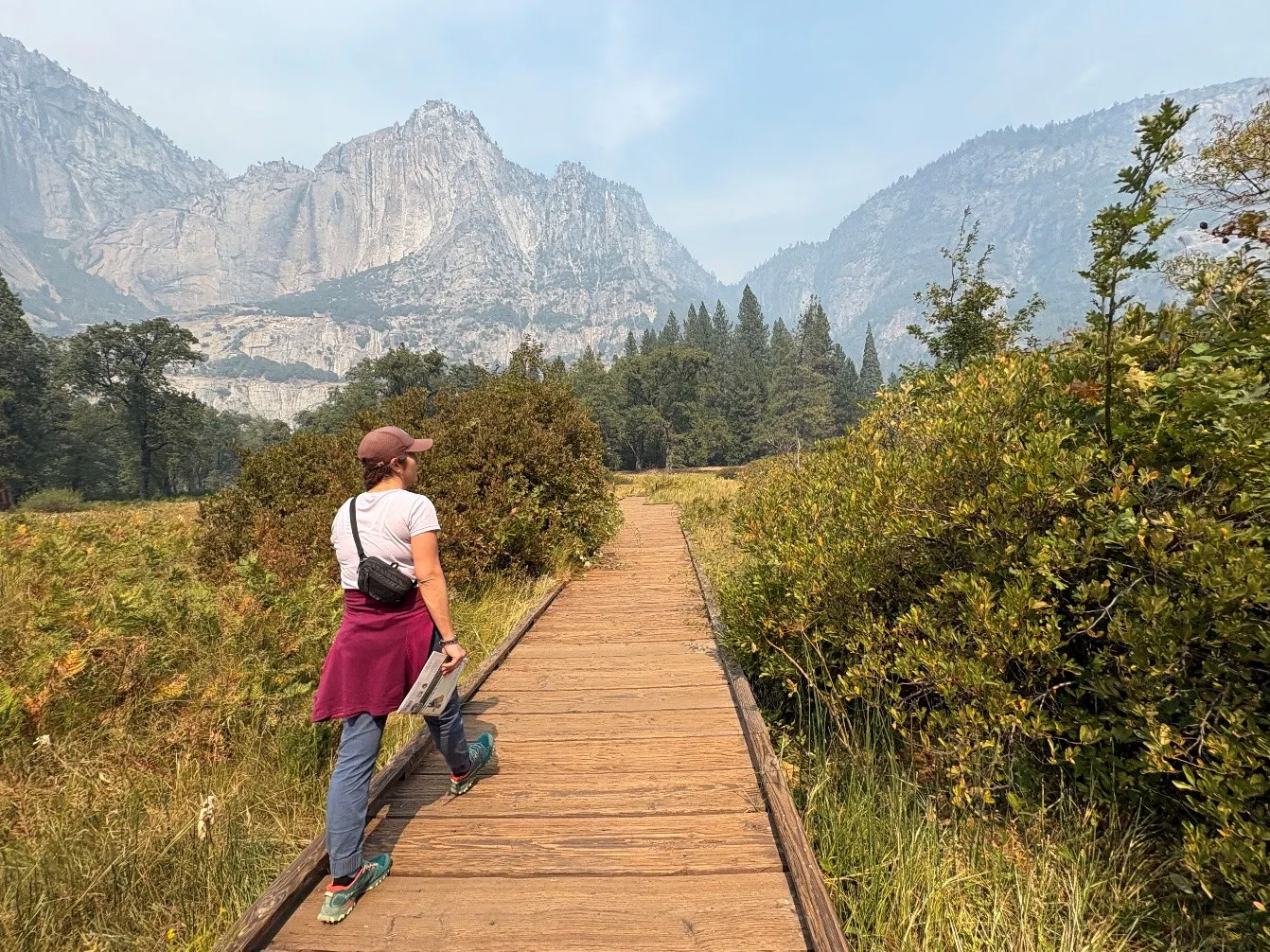 A person wearing a cap and backpack walks along a wooden boardwalk through a Yosemite meadow toward mountains.
