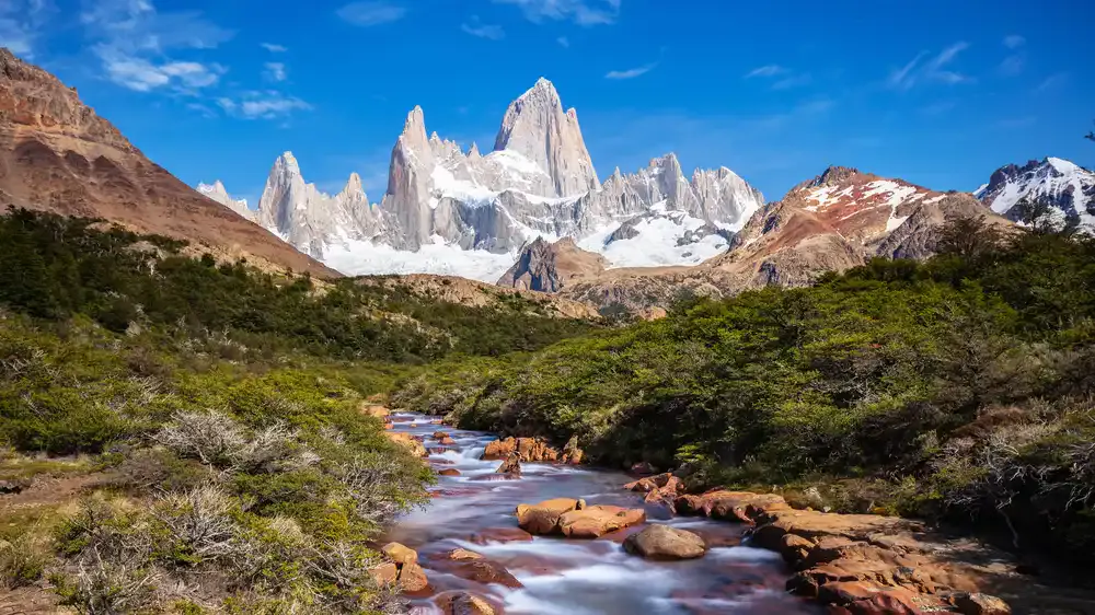 A clear stream flows through green vegetation called the Futaleufu River toward snow-capped jagged mountains under a bright blue sky.